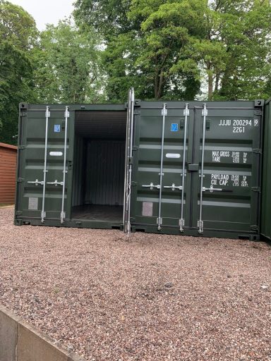 Green shipping container with one door open, placed on a gravel surface among trees.