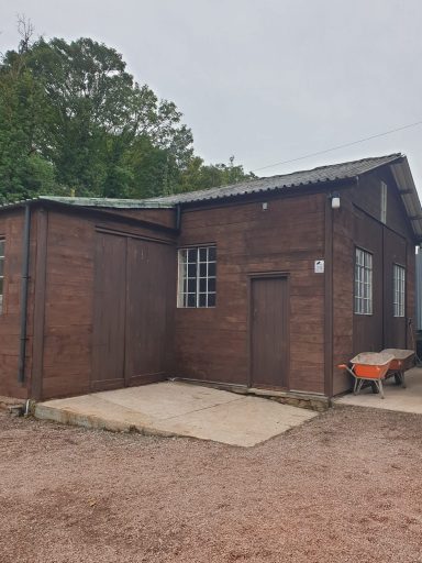 A small brown wooden building with a flat roof and a wheelbarrow nearby.